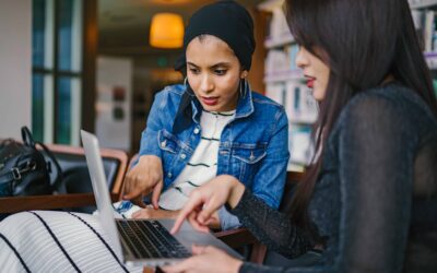 Two women in a library helping each other on a laptop.