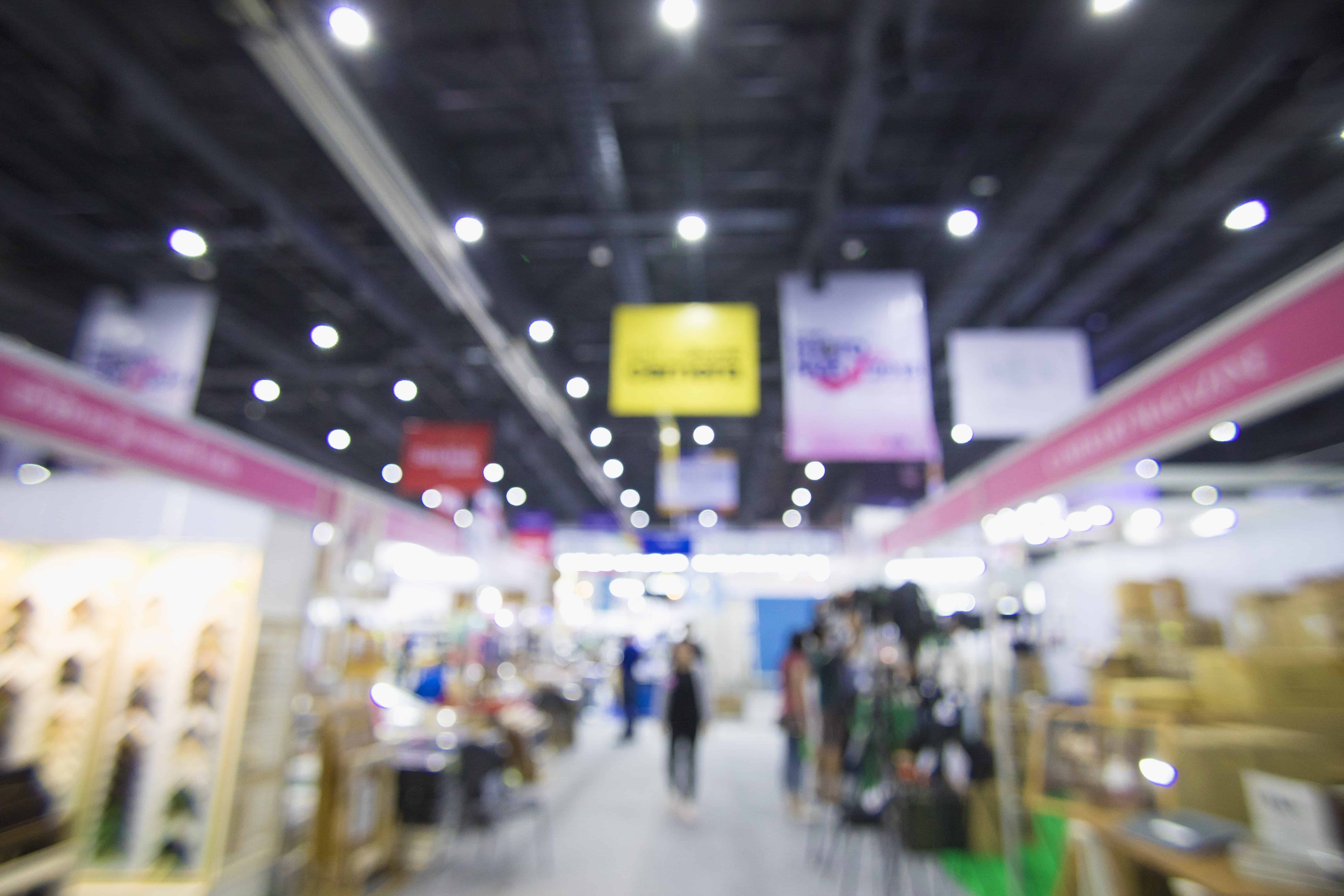 Blurred image of a trade show. Banners are clearly seen above booths in order to catch potential clients attention